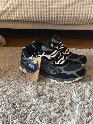 Asics NYC - Snygga svarta sneakers från Asics med grå och vita detaljer samt vita snören. Skorna har en sportig design med mesh och syntetmaterial för bra andningsförmåga. Ortholite-innersula för extra komfort. Perfekta för dig som gillar streetwear och sportig stil.