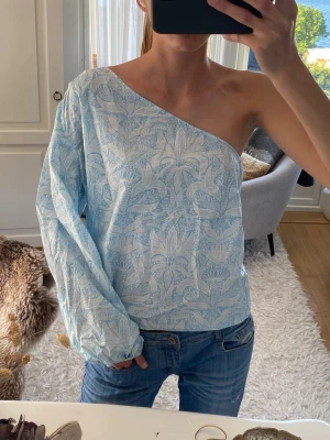 Ljusblå oneshoulder blus med mönster - Säljer en ljusblå blus med mönster och oneshoulder-design. Toppen har en lång ärm och en axel är bar, vilket ger en unik och trendig look. Tillverkad i ett lätt och luftigt material, perfekt för varmare dagar. Första bilden är min i storlek L passar även mindre, jag brukar bära S för referens☺️🌸