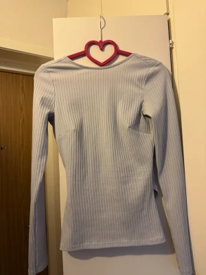 Gina Tricot topp ljusblå strl XS - Långärmad topp med omlottdesign framtill. Toppen har en figurnära passform och ribbad struktur. Den är designad med en V-ringning på ryggen som skapar en smickrande silhuett.