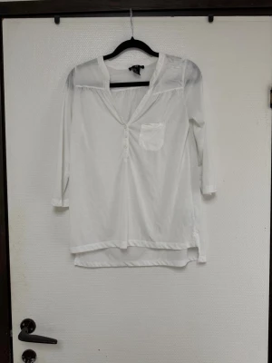 Vit blus med trekvartsärm från H&M - Stilren vit blus från H&M med trekvartsärm och v-ringning. Blusen har en liten bröstficka och knappar framtill. Tillverkad i ett tunt, lätt material som ger en avslappnad look. Perfekt att matcha med jeans eller kjol.