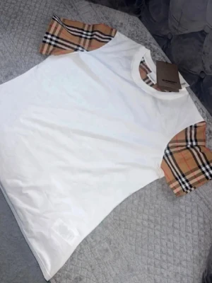  Burberry t-shirt  - Säljer en snygg vit t-shirt från Burberry med klassiska beige-rutiga korta ärmar. T-shirten har rund halsringning och är tillverkad i mjuk bomull. Perfekt för dig som vill ha en stilren men ändå unik look.