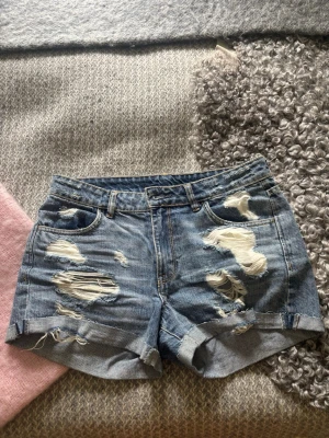 Blåa slitna jeansshorts med upprullad kant - Snygga blå jeansshorts med slitna detaljer och upprullad kant. Klassisk femficksmodell med bälteshällor och dragkedjegylf. Perfekta för en avslappnad och trendig sommarlook.