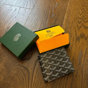 Svart plånbok med mönster - Snygg svart plånbok med det klassiska vita och gråa Goyardine-mönstret. Plånboken är vikbar och har diskret logga på framsidan. Kommer i original ask och med gul putsduk. Perfekt för dig som gillar exklusiva accessoarer.