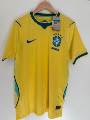 Brasilien fotbollströja Nike gul - Gul Brasilien fotbollströja från Nike med gröna detaljer och blå logga. Tröjan har CBF-emblem på bröstet, Dri-FIT-material och klassisk passform. Perfekt för dig som älskar fotboll och vill visa ditt stöd för Brasilien.