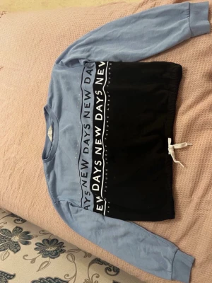 Blå och svart hoodie med text - Säljer en hoodie i blått och svart med trycket 'NEW DAYS' över bröstet. Tröjan har långa ärmar, ribbade muddar och en vit dragsko i nederkanten. Materialet känns mjukt och bekvämt, perfekt för chill dagar. Märket är ej synligt.