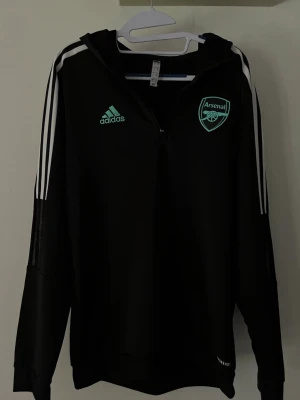 Svart Arsenal hoodie från Adidas - Cool svart hoodie från Adidas med Arsenal-logga och Adidas-märke i mintgrönt. Tröjan har dragkedja framtill, huva och klassiska vita ränder på ärmarna. Perfekt för fotbollsfans och sportiga dagar.