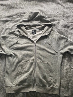 Grå zip-hoodie från Nike - Snygg grå hoodie från Nike med dragkedja och klassisk logga på bröstet. Tröjan har huva med dragsko och två stora fickor framtill. Mjuk insida och ribbade muddar vid ärmslut och nederkant. Perfekt för en avslappnad streetstyle.