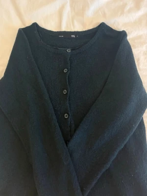 Svart kofta från 157 - Mysig mörk grön/blå kofta från 157 med rund halsringning och knäppning framtill. Koftan har en enkel och stilren design med långa ärmar och mjukt material som passar perfekt till lager-på-lager-stil.