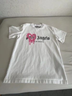 Palm angels tshirt - Säljer en vit t-shirt från Palm Angels med svart text 'Palm Angels Palm Beach' på bröstet och ett stort rosa hjärta med drip-effekt ovanpå. Klassisk passform och korta ärmar. Perfekt för dig som gillar streetwear och statement-plagg.