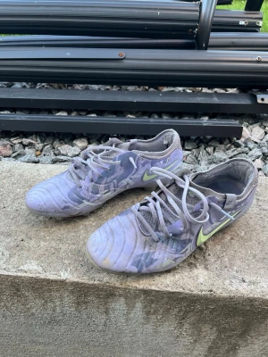 Lila Nike fotbollsskor med snörning - Säljer ett par lila Nike fotbollsskor med limegröna detaljer och snörning. Skorna har låg profil och är tillverkade i syntetmaterial med meshpartier för bra ventilation. Perfekta för dig som vill ha stilrena och lätta skor på planen.
