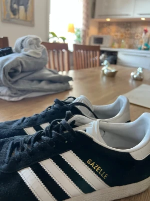 Adidas Gazelle svart/vit sneakers - Klassiska Adidas Gazelle sneakers i svart mocka med vita detaljer och de ikoniska tre ränderna på sidan. Låg modell med vit sula och guldfärgad Gazelle-text på sidan. Skön och tidlös sneaker som passar till det mesta.