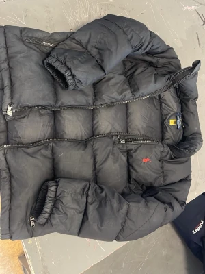 Svart pufferjacka från Polo Ralph Lauren - Snygg svart pufferjacka från Polo Ralph Lauren med röd broderad logga på bröstet. Jackan har dragkedja framtill, två sidofickor med dragkedja och elastiska muddar vid ärmarna. Perfekt för kalla vinterdagar och riktigt trendig stil. Helt original 