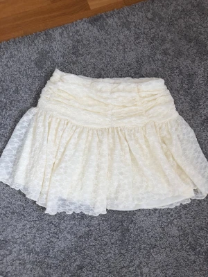 Vit spetskjol med volang - Söt kort kjol i vit spets med volang och blommigt mönster. Kjolen har en hög midja och är fodrad under spetsen för extra komfort. Perfekt för dig som gillar romantisk och feminin stil.