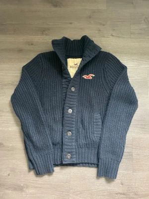 Mörkblå stickad kofta Hollister - Mysig mörkblå stickad kofta från Hollister med grov ribbstickning, stora knappar framtill och två fickor. Koftan har hög krage och Hollister-logga broderad på bröstet. Perfekt för lager på lager och chill stil.