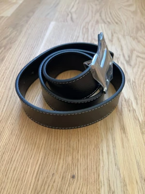 Svart skärp med silverfärgat H-spänne - Stilrent svart bälte med vit kontrastsöm och ett stort, silverfärgat H-format spänne. Bältet har en klassisk design och är tillverkat i skinn, vilket ger en lyxig känsla. Perfekt accessoar för att lyfta din outfit.