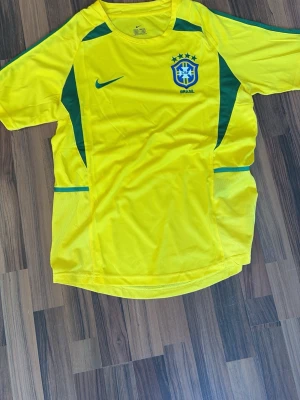 Brasilien hemma tröja 2002 - Snygg gul fotbollströja från Nike med Brasilien-emblem och gröna detaljer. Tröjan har korta ärmar, rund hals och är tillverkad i ett lätt, ventilerande material som passar perfekt till träning eller match. Klassisk design med Nike-logga på bröstet. Perfekt för personer som gillar retro tröjor 