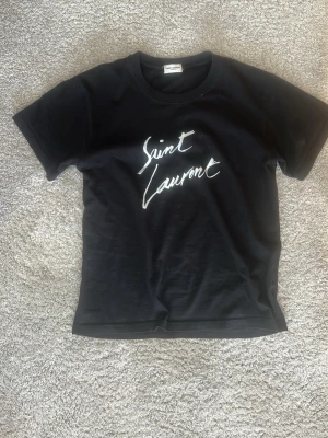 Svart Saint Laurent t-shirt - Snygg svart t-shirt från Saint Laurent med vit logga på bröstet. Klassisk rund halsringning och korta ärmar. Perfekt för dig som gillar stilrena och ikoniska plagg. 