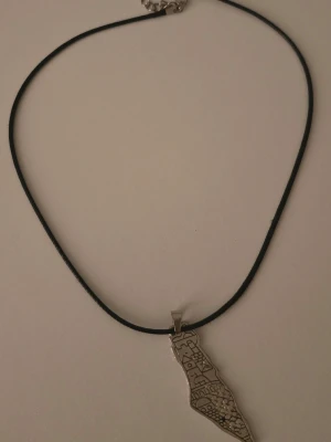 Halsband med karta i silverfärg - Unikt halsband med svart snöre och ett silverfärgat hänge i form av en karta med ingraverade mönster. Hänget har en avlång, oregelbunden form och detaljerade linjer som ger en cool och personlig touch. Perfekt accessoar för dig som gillar statement-smycken.