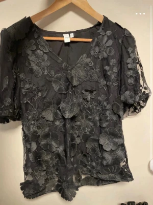 Svart meshblus med blommig spets - Säljer en svart transparent meshblus från YAS  med korta puffärmar och detaljerade blommiga spetsapplikationer över hela plagget. 