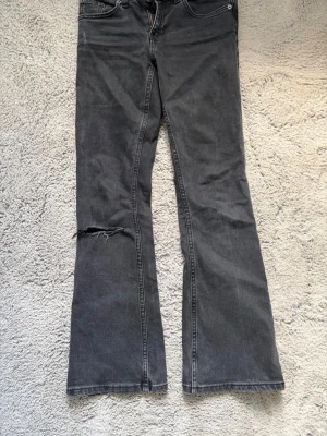 Mörkgrå bootcut jeans  - Säljer ett par mörkgrå bootcut jeans med slitning på ena benet. Jeansen har klassisk femficksdesign och normal passform upptill med utsvängda ben. Perfekta för dig som gillar en avslappnad och trendig look.
