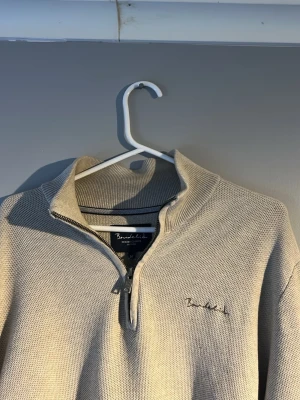 Beige stickad halfzip  - Snygg beige stickad med halv dragkedja och hög krage. Tröjan har diskret broderad logga på bröstet och är gjord i mjukt material som känns skönt mot huden. Perfekt till jeans eller chinos.