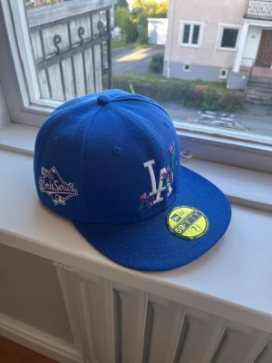 Blå LA Dodgers keps från New Era - Snygg blå snapback-keps från New Era med LA Dodgers-logga framtill och broderade blommor. Kepsen har platt skärm, broderad North Series-patch på sidan och klassisk New Era-logga. Perfekt för dig som gillar streetwear och sportig stil.