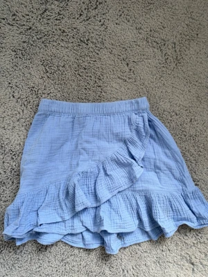 Ljusblå volangkjol från Gina Tricot - Supersöt ljusblå kortkjol från Gina Tricot med volangdetaljer och omlottkänsla. Kjolen har resår i midjan och är gjord i ett mjukt, luftigt bomullstyg som passar perfekt till varmare dagar. Enkel att matcha med både toppar och sneakers.