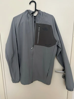 Grå vindjacka från The North Face - Snygg grå vindjacka från The North Face med hel dragkedja, huva och en mörkare bröstficka med dragkedja. Jackan har elastiska muddar vid ärmsluten och är tillverkad i ett lätt, vindtätt material. Perfekt för blåsiga dagar.