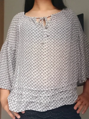 Vit mönstrad blus med knytning - Luftig vit blus med svart småmönster och rund hals med knytning. Blusen har trekvartsärm och ett lätt transparent material som ger en avslappnad look. Passar perfekt till jeans eller shorts för en cool stil.