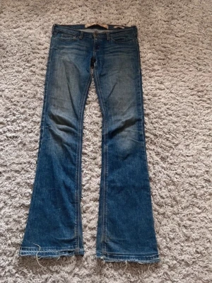 Hollister Jeans - Säljer ett par blå bootcut jeans med råa, fransiga benslut och klassisk femficksdesign. Jeansen har en snygg tvätt med lätt slitning på låren och bakfickor med dekorativa sömmar. Perfekta för dig som gillar retrovibbar och avslappnad stil. Midjemåttet är 39,5 cm rakt över och innerbenslängden är 81 cm.