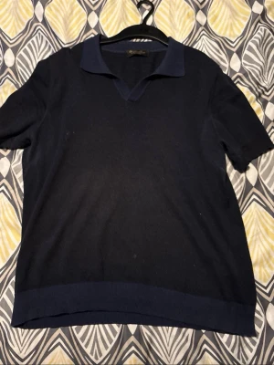 Mörkblå cashmerepiké från Loro Piana - Exklusiv mörkblå stickad pikétröja från Loro Piana i 100% cashmere. Tröjan har korta ärmar, klassisk krage och ribbstickad nederkant. Tillverkad i Italien och perfekt för dig som gillar lyxiga material och stilren design. Solblekt, kvitto finns! 