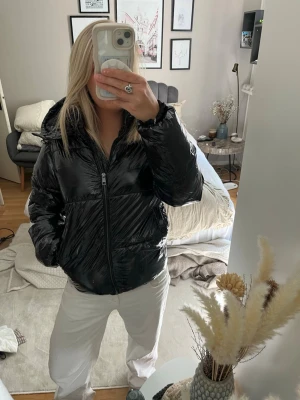 Svart glansig pufferjacka med huva - Säljer en svart glansig pufferjacka med dragkedja och huva. Jackan har en oversized look och två sidofickor. Perfekt för kalla dagar när du vill hålla dig varm och ändå se cool ut.