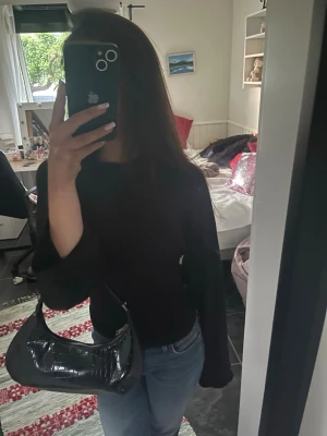 Svart axelväska i krokodil material - Snygg svart axelväska i krokodil material med kort rem. Väskan har en stilren, halvmåneformad design och blanka detaljer. Perfekt för dig som vill ha en trendig och enkel accessoar till din outfit.
