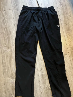 Svarta träningsbyxor från Reebok - Säljer ett par svarta träningsbyxor från Reebok med elastisk midja och diskret logga på benet. Byxorna är tillverkade i ett lätt och smidigt material som passar perfekt för träning eller sportaktiviteter.