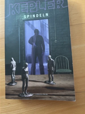 Spindeln av Lars Kepler - En spännande pocketbok, 'Spindeln' av Lars Kepler. Omslaget är mörkt med lila och svarta toner, och visar en mystisk figur i en dörröppning samt små statyetter på ett bord. Perfekt för dig som gillar nervkittlande thrillers och deckare.