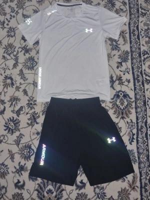 Under Armour träningsset grå/svart - Snyggt träningsset från Under Armour med en ljusgrå t-shirt och svarta shorts. T-shirten har tryckta loggor och textdetaljer, rund hals och korta ärmar. Shortsen har elastisk midja och reflekterande logga samt texten 'ARMOUR' på benet. Perfekt för gym eller löpning.