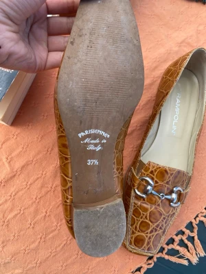 Bruna loafers i krokopräglat läder - Snygga bruna loafers från Trampolini i krokopräglat läder med metallspänne framtill. Skorna har en låg klack och fyrkantig tå, tillverkade i Italien. Perfekta för dig som vill ha en unik och trendig look.