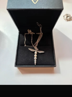 Maria Nilsdotter Silverhalsband med pärlor och vingar - Säljer mitt Maria Nilsdotter halsband så fint längd 50 cm! Alla kvitton alla boxar och påsar och allt tillkommer!Unikt halsband i silver med ett hängsmycke format som en trollslända. Kroppen består av fyra vita pärlor och ett spetsigt avslut, medan vingarna är detaljerat utformade i metall. Kedjan är tunn och elegant, perfekt för dig som gillar naturinspirerade smycken. Nypris 3300
