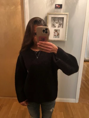 Svart oversized stickad tröja - Mysig svart stickad tröja med oversized passform och långa ärmar. Tröjan har en enkel rund halsringning och är tillverkad i ett fluffigt, mjukt material som ger en avslappnad look. Perfekt för kalla dagar och enkel att matcha med jeans.