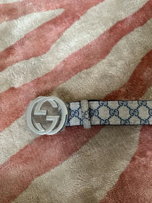 GG beige och blått bälte - Säljer ett ikoniskt gg-bälte med GG-monogram i blått på beige bakgrund. Bältet har ett stort, runt spänne i silverfärgad metall med klassiska GG-loggan. Perfekt accessoar för att lyfta vilken outfit som helst.