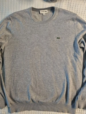 Grå rundhalsad tröja från Lacoste - Snygg ljusgrå tröja från Lacoste med klassisk rund halsringning och den ikoniska gröna krokodilloggan broderad på bröstet. Tröjan har ribbade muddar vid ärmslut och nederkant. Litet hål i ena armhålan, se bild. Därav priset. Fast pris, inga byten.