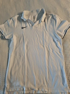 Vit pikétröja från Nike  - Vit pikétröja från Nike med klassisk krage och två knappar framtill. Diskret svart Nike-logga på bröstet och svarta detaljer på kragen och ärmslut. Perfekt för dig som gillar sportig stil och enkel design.