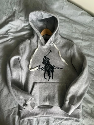 Grå hoodie från Ralph Lauren - Snygg grå hoodie från Ralph Lauren med stor svart polospelare och logga på bröstet. Hoodien har vita dragsnören, stor känguruficka och ribbade muddar. Perfekt för en chill och stilren look. Storlek M men är kort på mig som är 190 lång så passar bättre som S