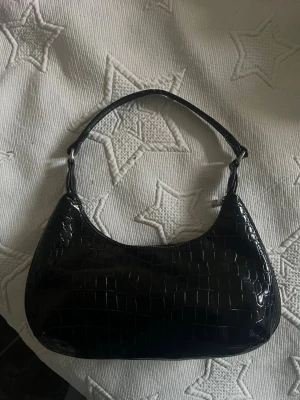 Svart axelväska i krokodil material - Snygg svart axelväska i krokodil material med kort rem. Väskan har en stilren, halvmåneformad design och blanka detaljer. Perfekt för dig som vill ha en trendig och enkel accessoar till din outfit.