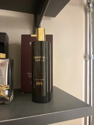Zara Immortal Vanilla parfym 🤎 - En elegant parfymflaska från Zara med doften Immortal Vanilla. Flaskan är cylinderformad i svart glas med guldfärgad text och ett matchande guldfärgat lock. Perfekt för dig som gillar lyxiga och moderna dofter med en touch av vanilj.