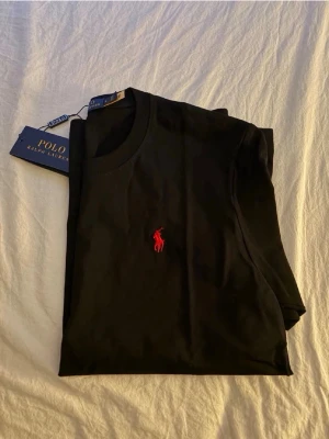 Svart t-shirt från Polo Ralph Lauren - Klassisk svart t-shirt från Polo Ralph Lauren med röd broderad logga på bröstet. T-shirten har rund hals och är kortärmad, perfekt till jeans eller shorts. Tillverkad i mjuk bomull för skön känsla hela dagen. Pris kan diskuteras 