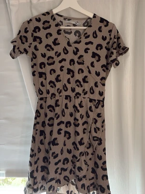 Leopardmönstrad klänning Lager 157 - Säljer en beige omlottklänning från lager 157 med svart leopardmönster. Klänningen har korta ärmar, markerad midja och volangkant nedtill i barnstorlek 160.