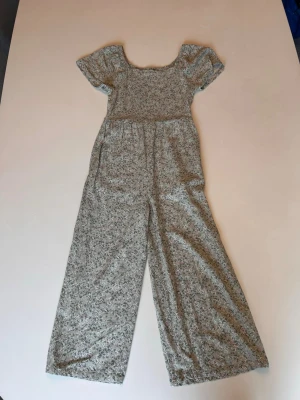 Blommig jumpsuit med vida ben storlek 9-10 - Supersöt jumpsuit med småblommigt mönster i grönt och vitt. Vida ben och korta puffärmar ger en avslappnad vibe. Smockad överdel och fyrkantig ringning. Perfekt för dig som gillar en romantisk och trendig stil.