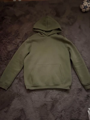 Olivgrön hoodie med magficka - Extremt skön och bra kvalitet. En stilren olivgrön hoodie med klassisk huva och stor magficka framtill. Hoodien har ribbade muddar vid ärmslut och nederkant. Perfekt för en avslappnad och trendig look. Materialet känns mjukt och bekvämt mot huden.