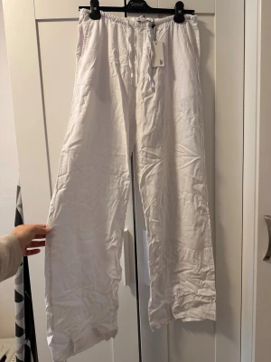 Vita linnebyxor med resårmidja - Säljer ett par vita linnebyxor med avslappnad passform och resår i midjan. Byxorna har raka ben och är tillverkade i ett lätt och luftigt linnematerial, perfekta för en chill och stilren look.
